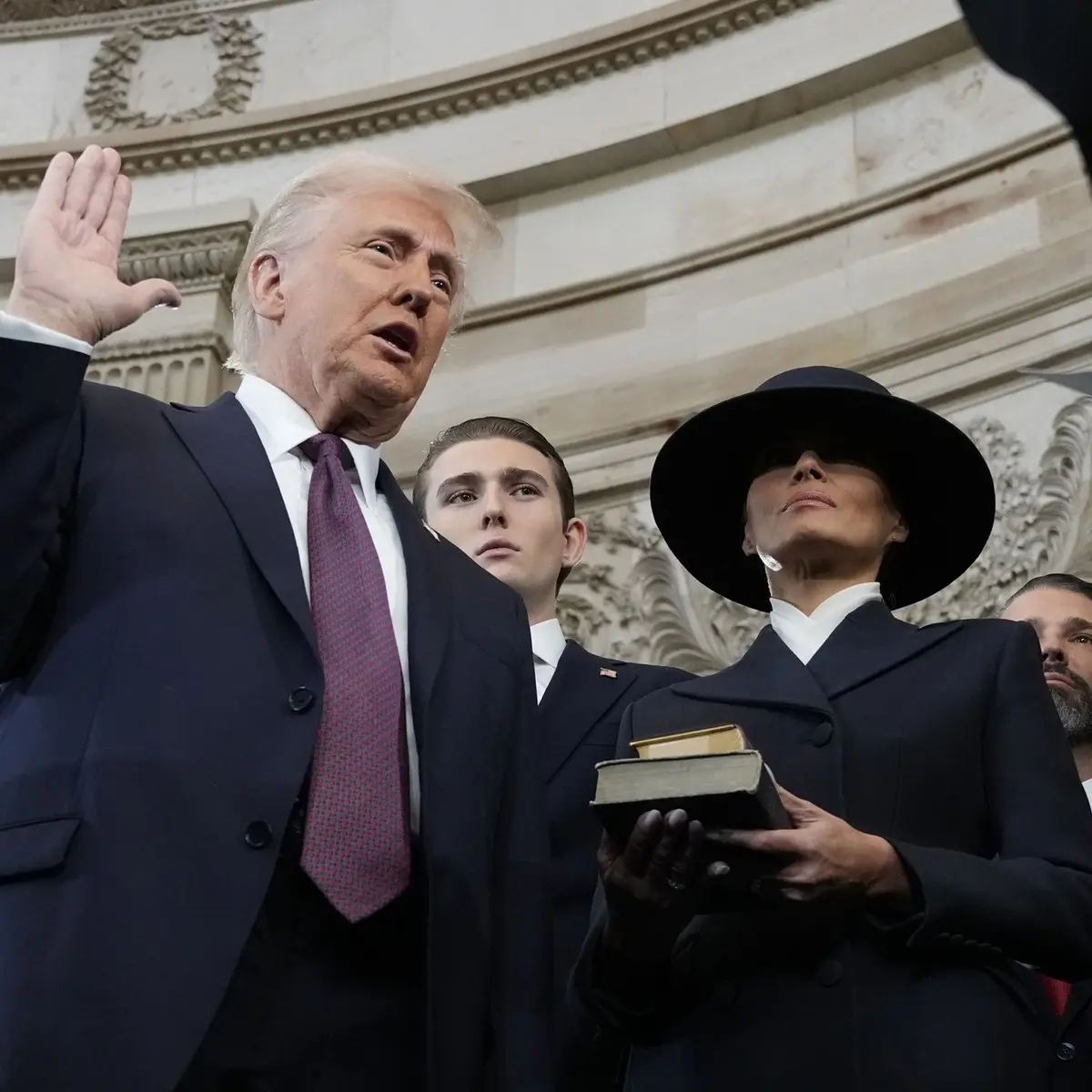 a low-angle photo shows Donald Trump raising his right hand while taking an oath of office, with Melania Trump holding a…