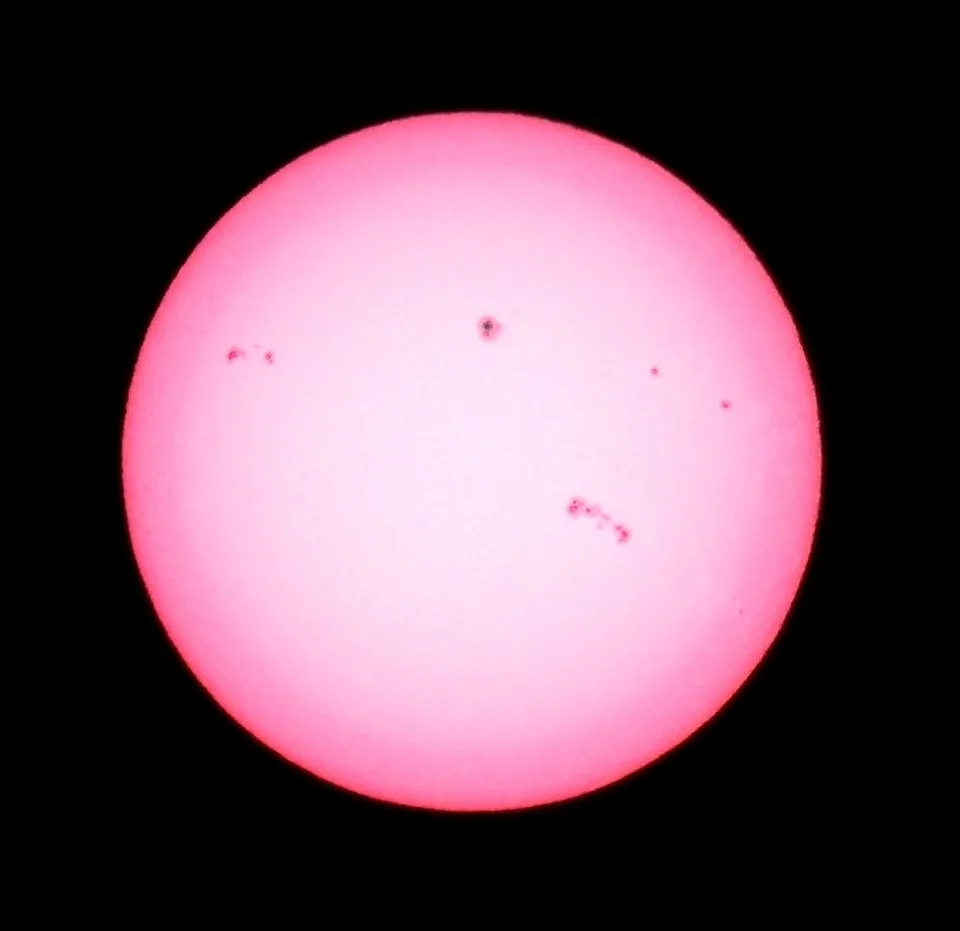 A pink-hued, circular image of the sun against a black background, with several small dark spots visible on its surface…