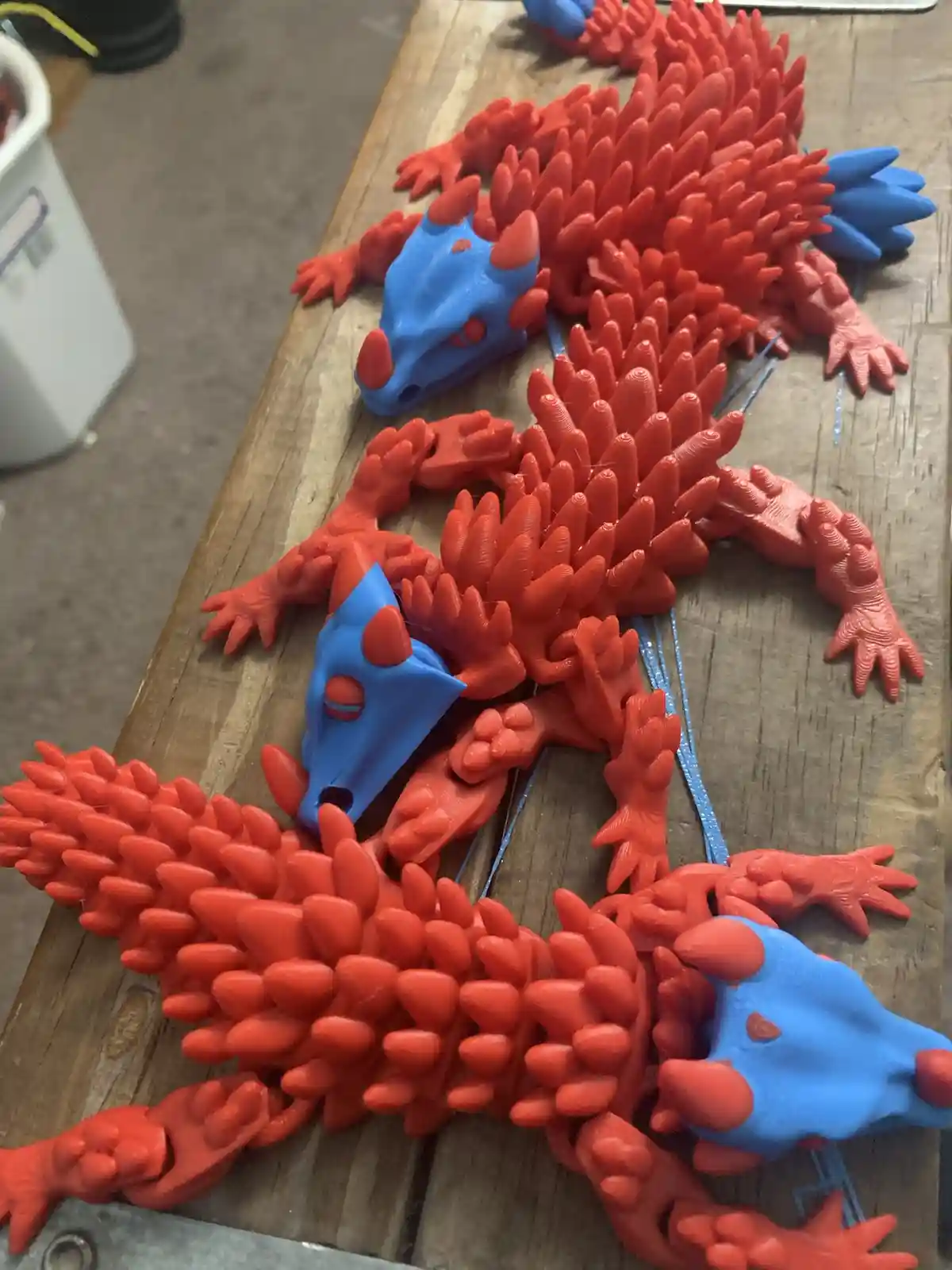 Several 3d-printed dragon figurines, mostly red with blue heads, are arranged on a wooden surface. The dragons have spiky…
