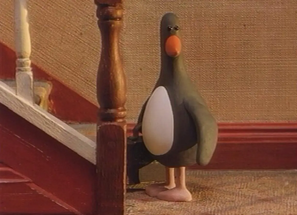 A stop-motion animated penguin with a black body, white belly, orange beak, and pink feet stands at the bottom of a wooden…