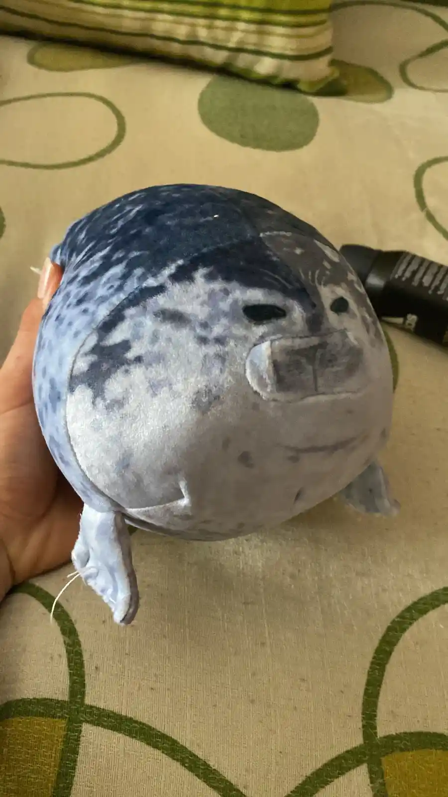 A person holds a round, blue-and-gray spotted plush seal toy with a stitched face, against a patterned green and beige…