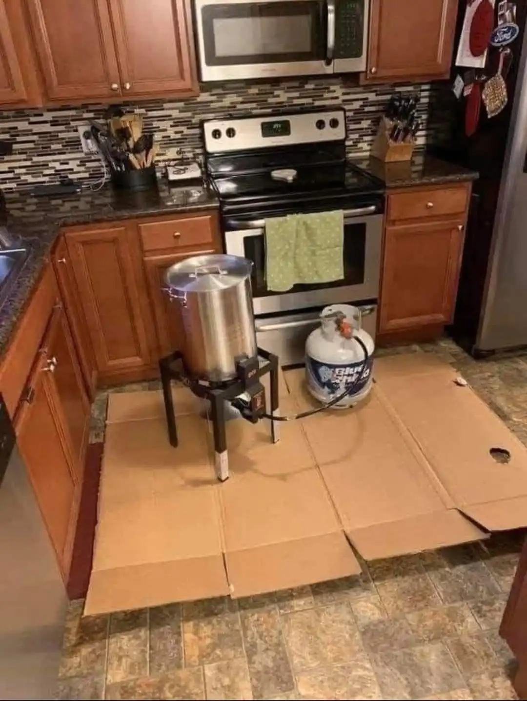 A kitchen scene shows a large metal pot on a portable burner connected to a propane tank, both placed on a large flattened…