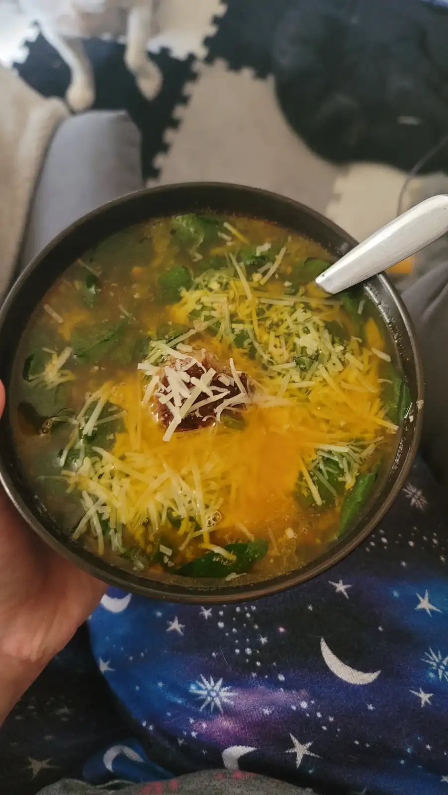 A hand holds a dark bowl of soup with a spoon, topped with shredded yellow and white cheese and green leafy vegetables. The…