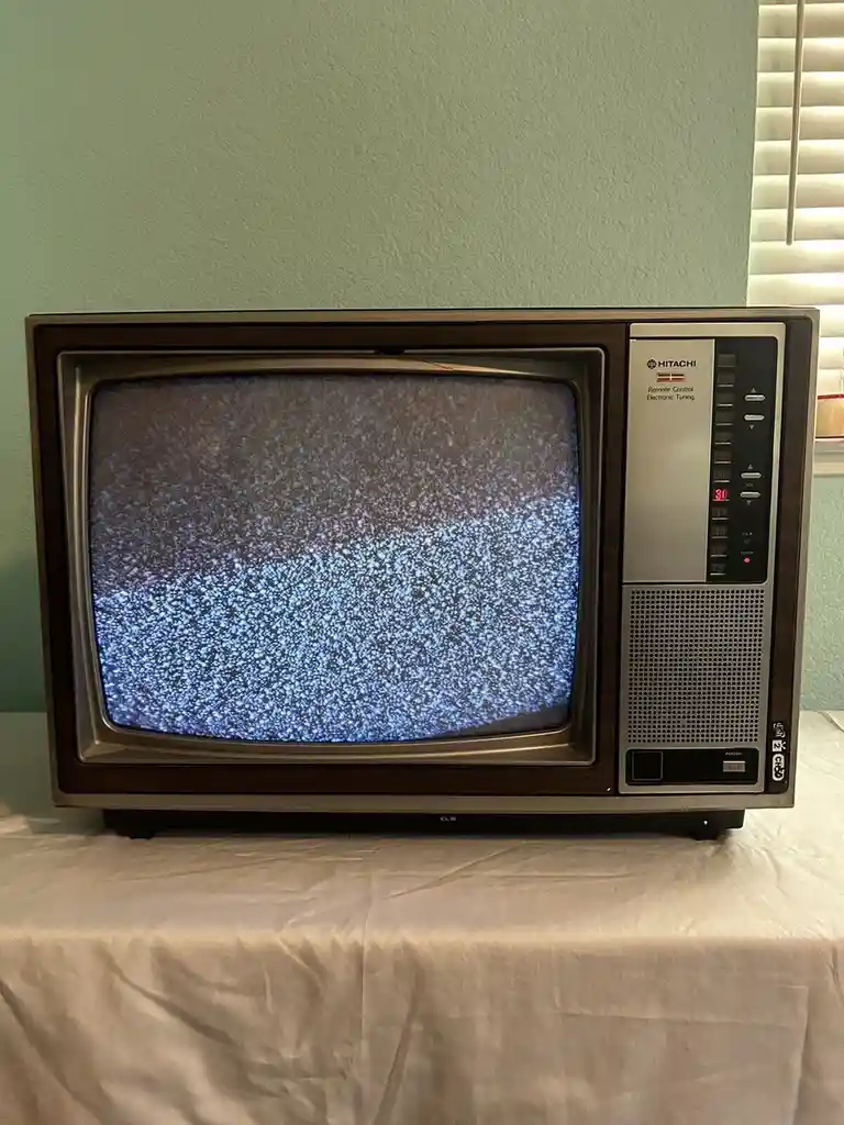 An old Hitachi CRT television sits on a white cloth, displaying static noise on its screen. The right side panel shows…