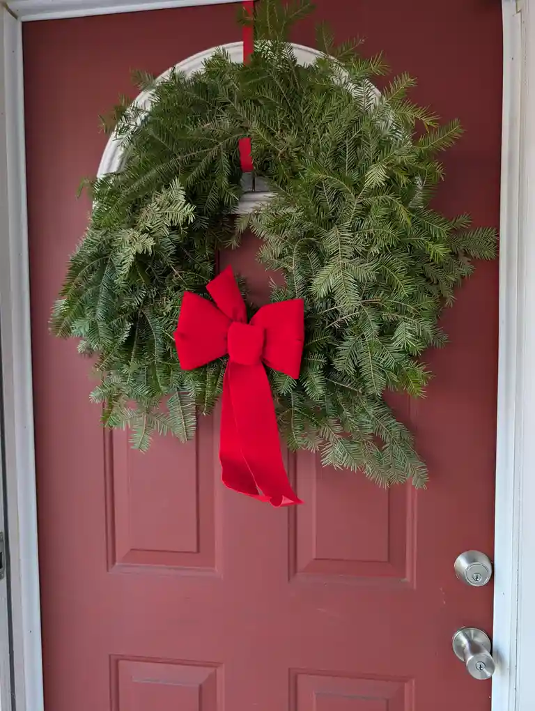 A large, lush green pine wreath with a prominent red bow hangs on a red door, contrasting with the door's color. The…