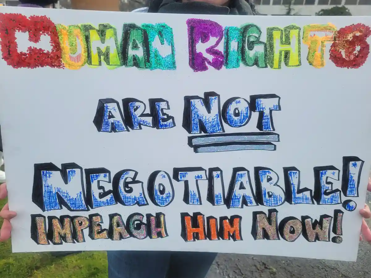 A person holds a handmade protest sign with colorful, glittery, and scribbled text. The top line reads 'HUMAN RIGHTS' in…