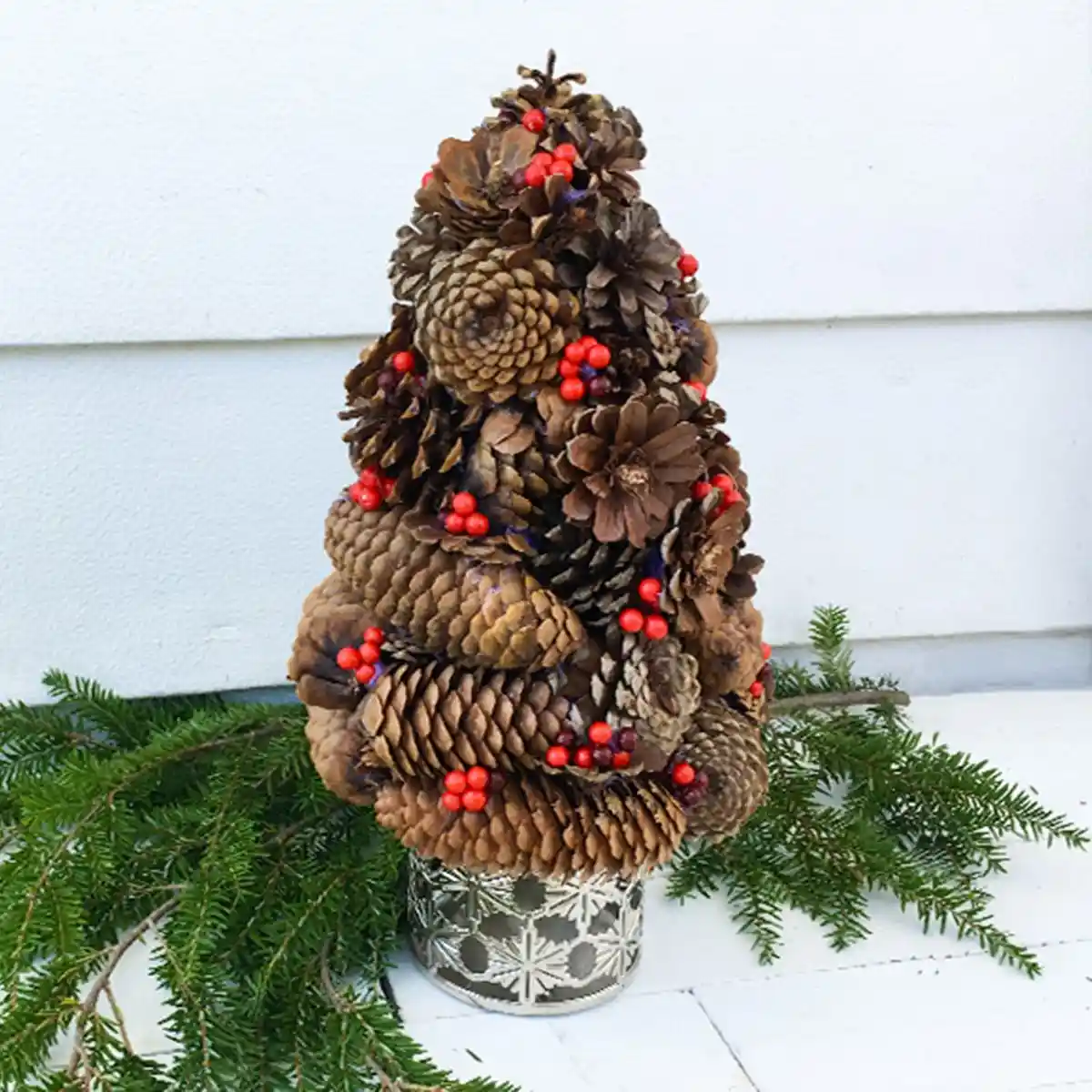 A decorative Christmas tree made entirely of brown pinecones, adorned with small red berries, sits in a silver…