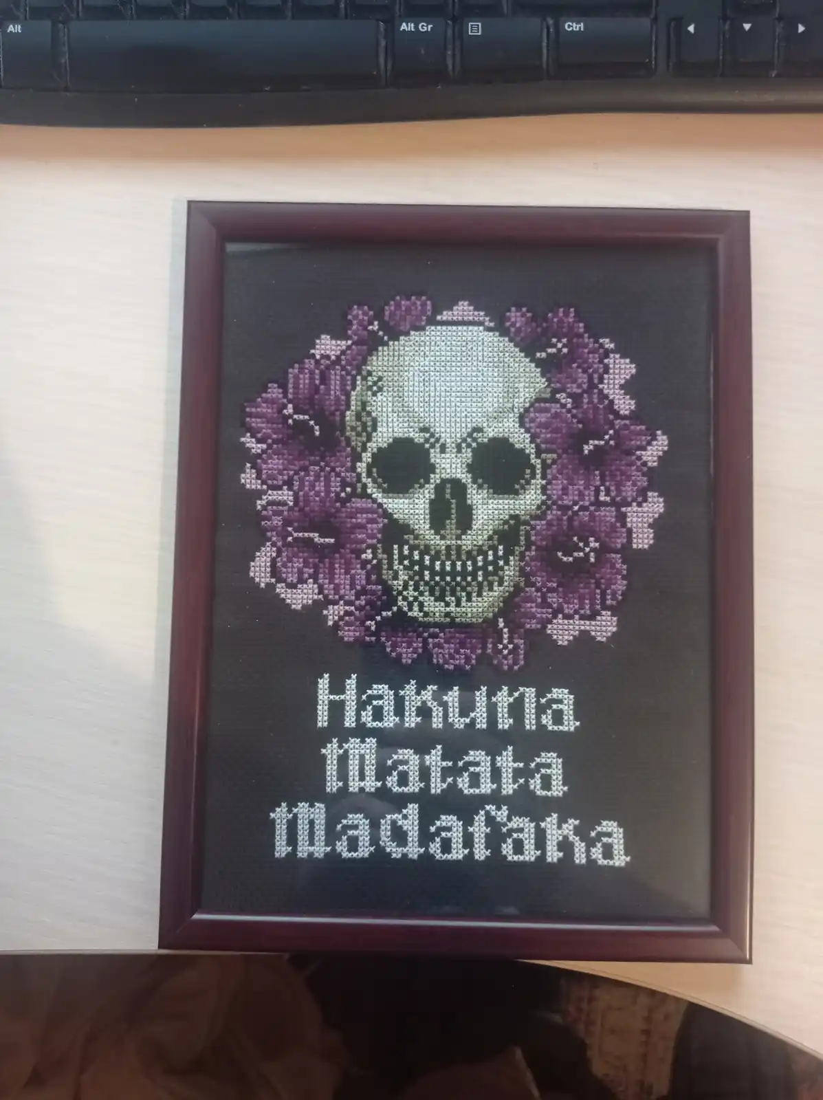 A framed cross-stitch artwork on a wooden desk shows a skull surrounded by purple flowers, with the text 'Hakuna Matata…