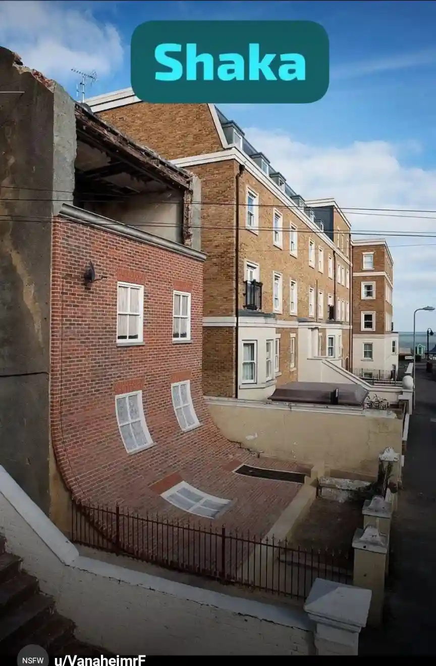 A photograph of a brick building with a severely warped, curved facade, making it look like a distorted face with windows as…