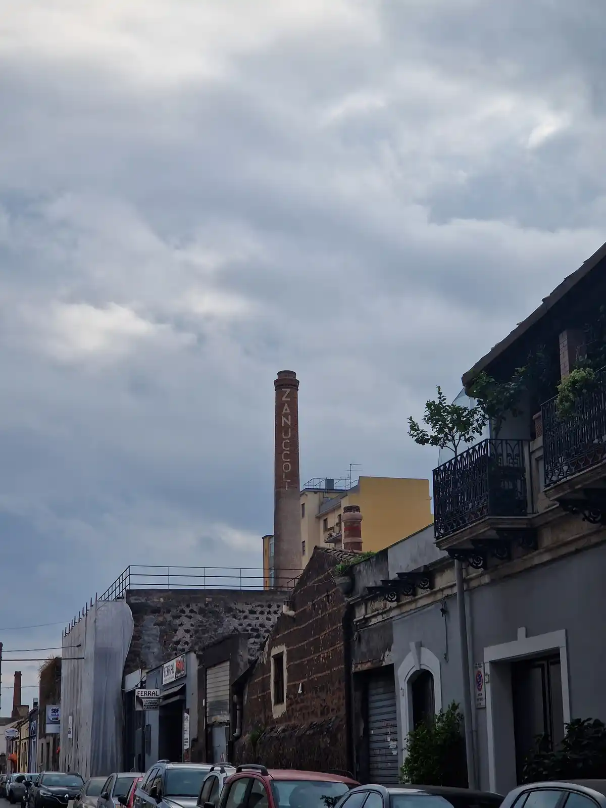 A street view shows a tall brick chimney with the word 'ZANUCCO' written vertically on its side, rising above old buildings…