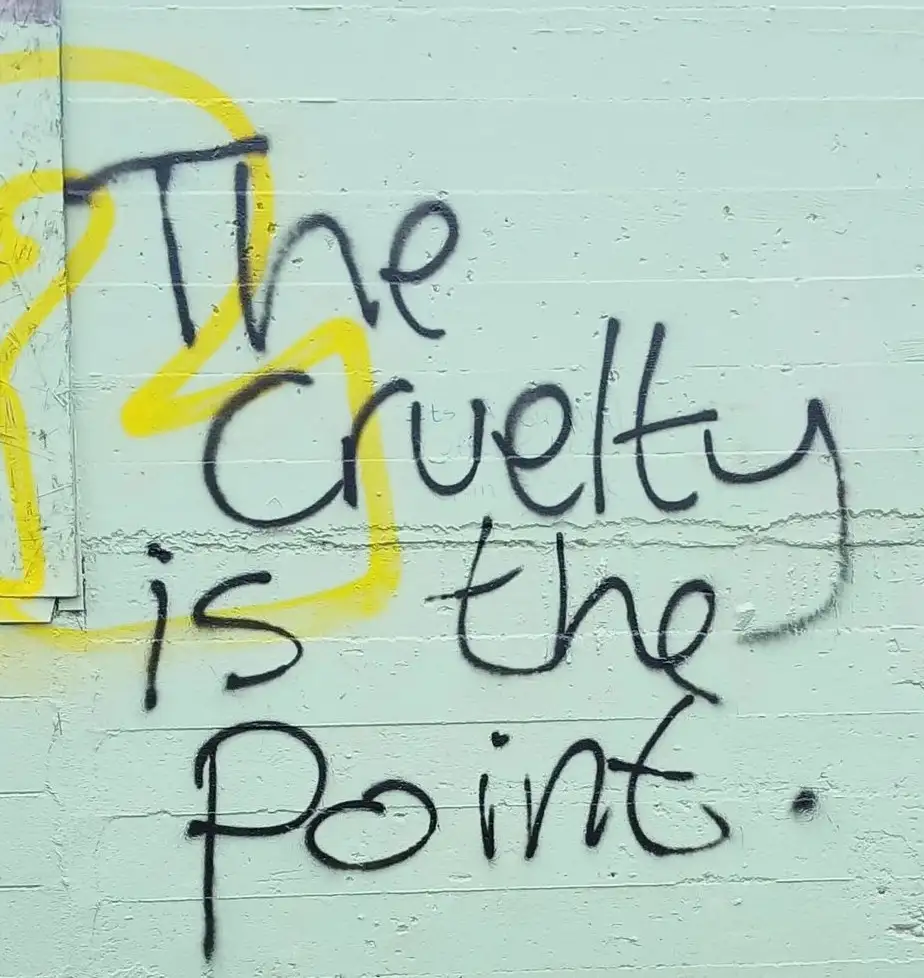 A close-up of graffiti on a light green painted brick wall, featuring the phrase 'The Cruelty is the Point.' written in…