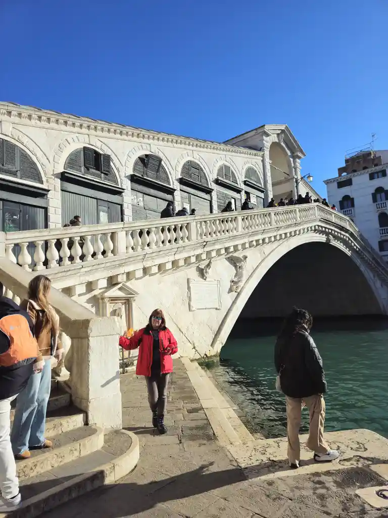A woman in a red jacket and sunglasses stands on a stone walkway beside the iconic stone arch bridge in Venice, Italy, under…