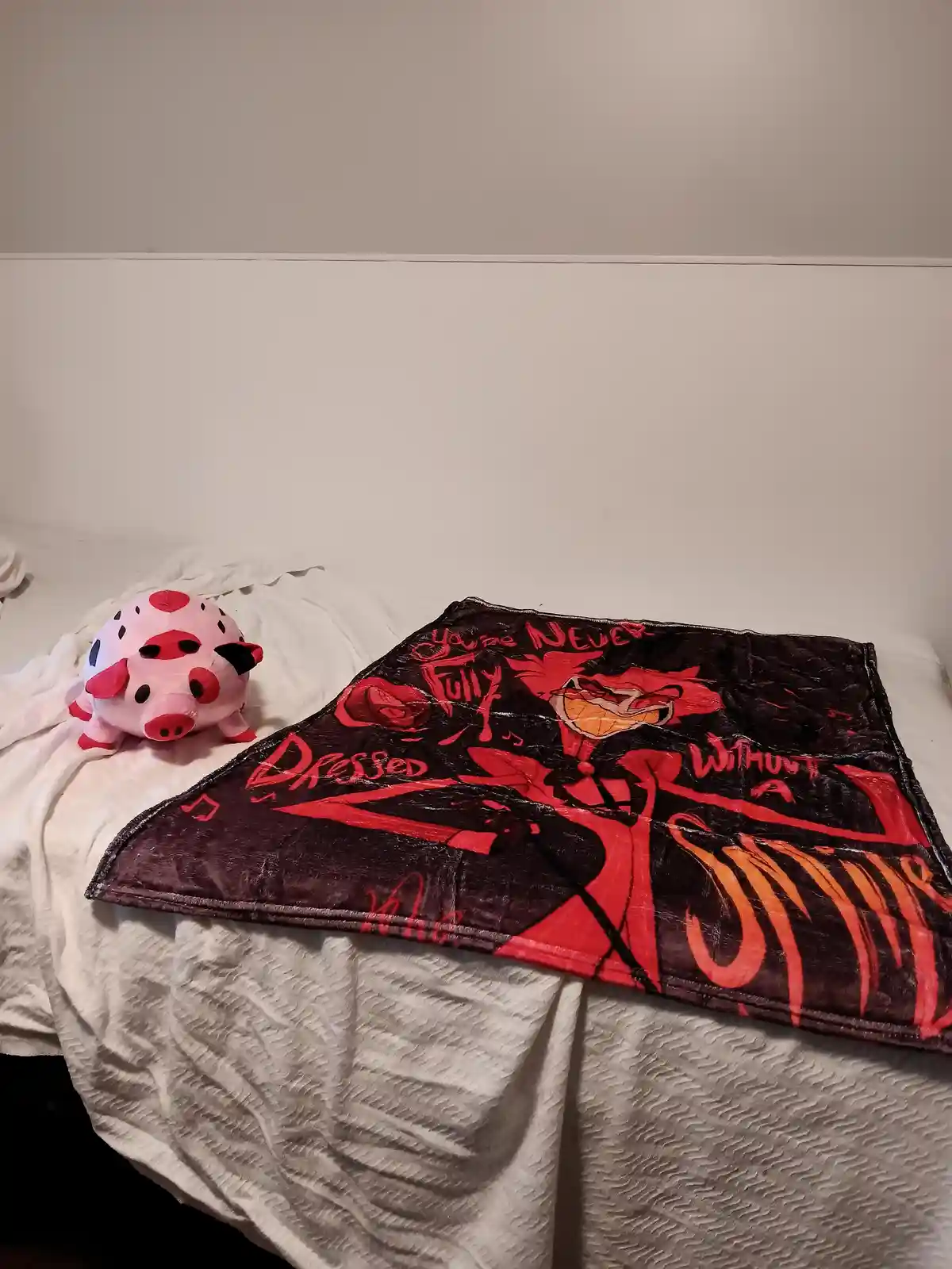 A white bed has a pink plush pig toy on the left and a dark red blanket with a cartoon character and text on the right. The…