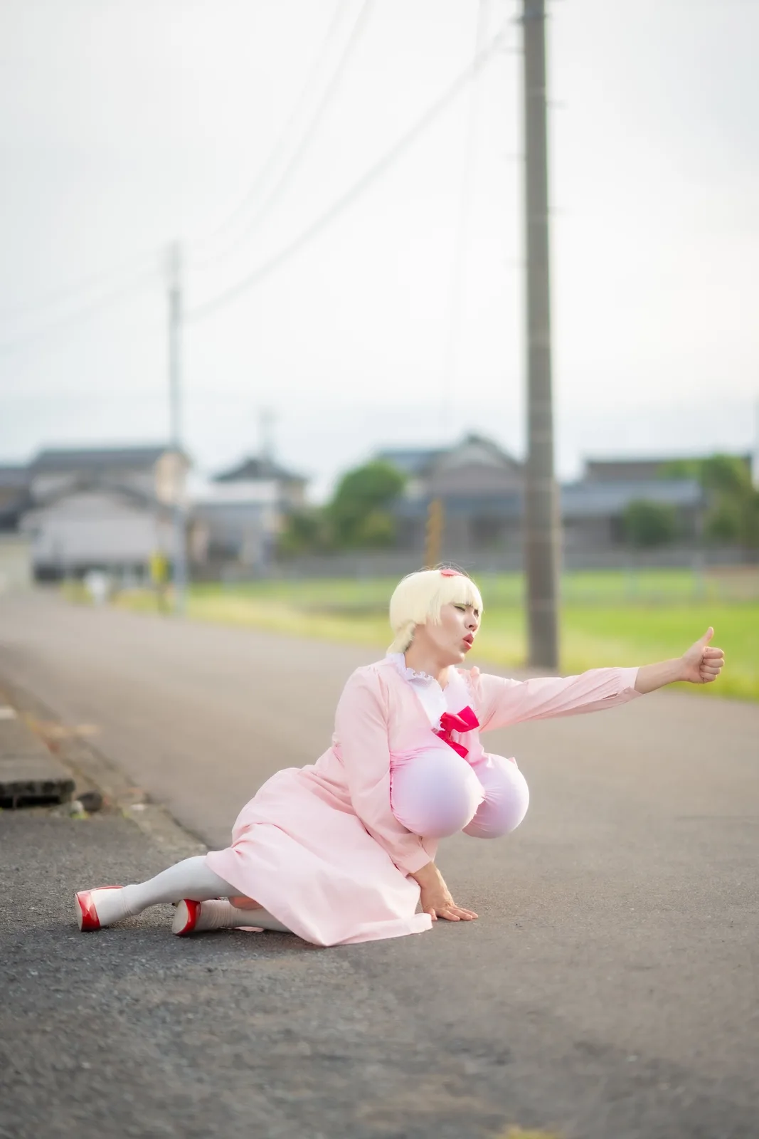 A person in a pink outfit with blonde wig and red shoes is crouching on an asphalt road, giving a thumbs-up while puckering…