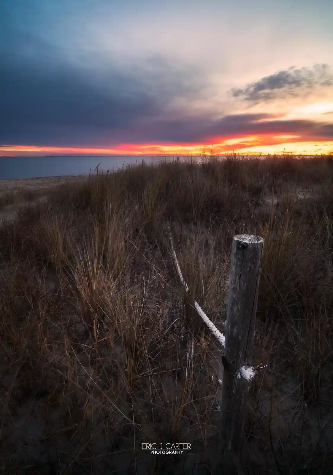A serene beach sunset photo featuring tall brown dune grass, a weathered wooden post with a white rope, and a dramatic sky…