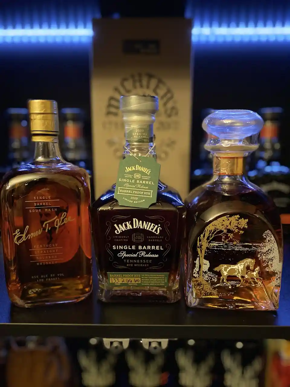 Three premium whiskey bottles are displayed on a shelf under blue lighting: Elmer T. Lee Single Barrel Sour Mash Bourbon, a…