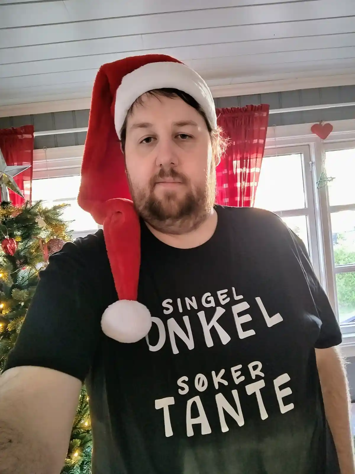 A man with a beard wears a red Santa hat and a black t-shirt with white text that reads 'SINGEL ONKEL SØKER TANTE'. He…