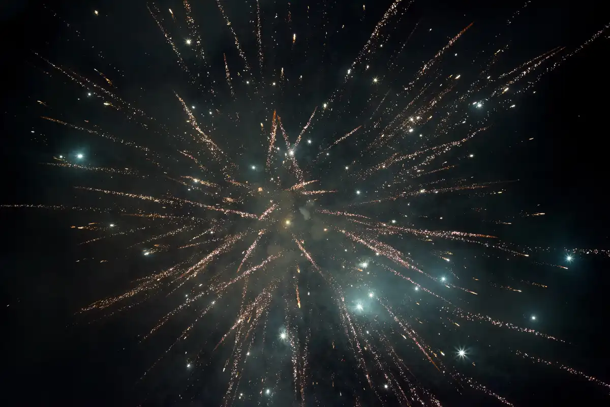 A vibrant nighttime photograph captures a massive fireworks explosion, with golden-orange and white sparks radiating outward…