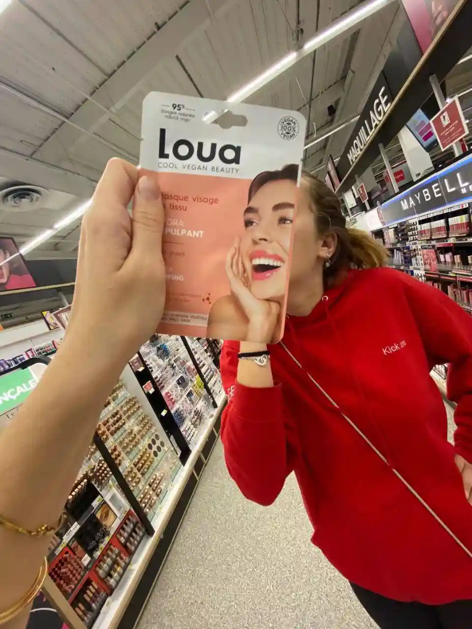 A person in a red hoodie holds up a Loua brand facial sheet mask package in a cosmetics aisle, with the product's smiling…