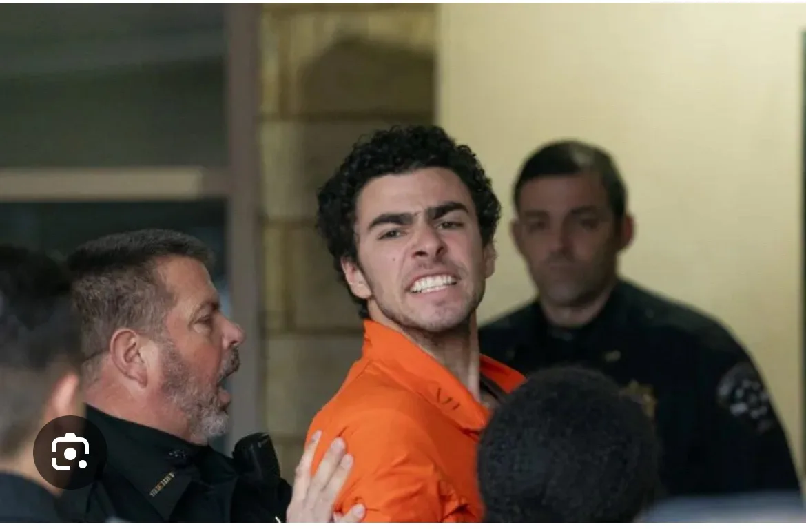A man in an orange prison jumpsuit is being escorted by police officers, his face contorted in an angry, yelling expression…