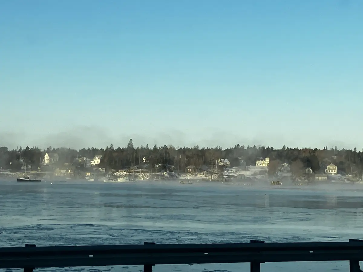 A scenic winter view from a roadside, showing a frozen body of water with mist rising from its surface, across which a…