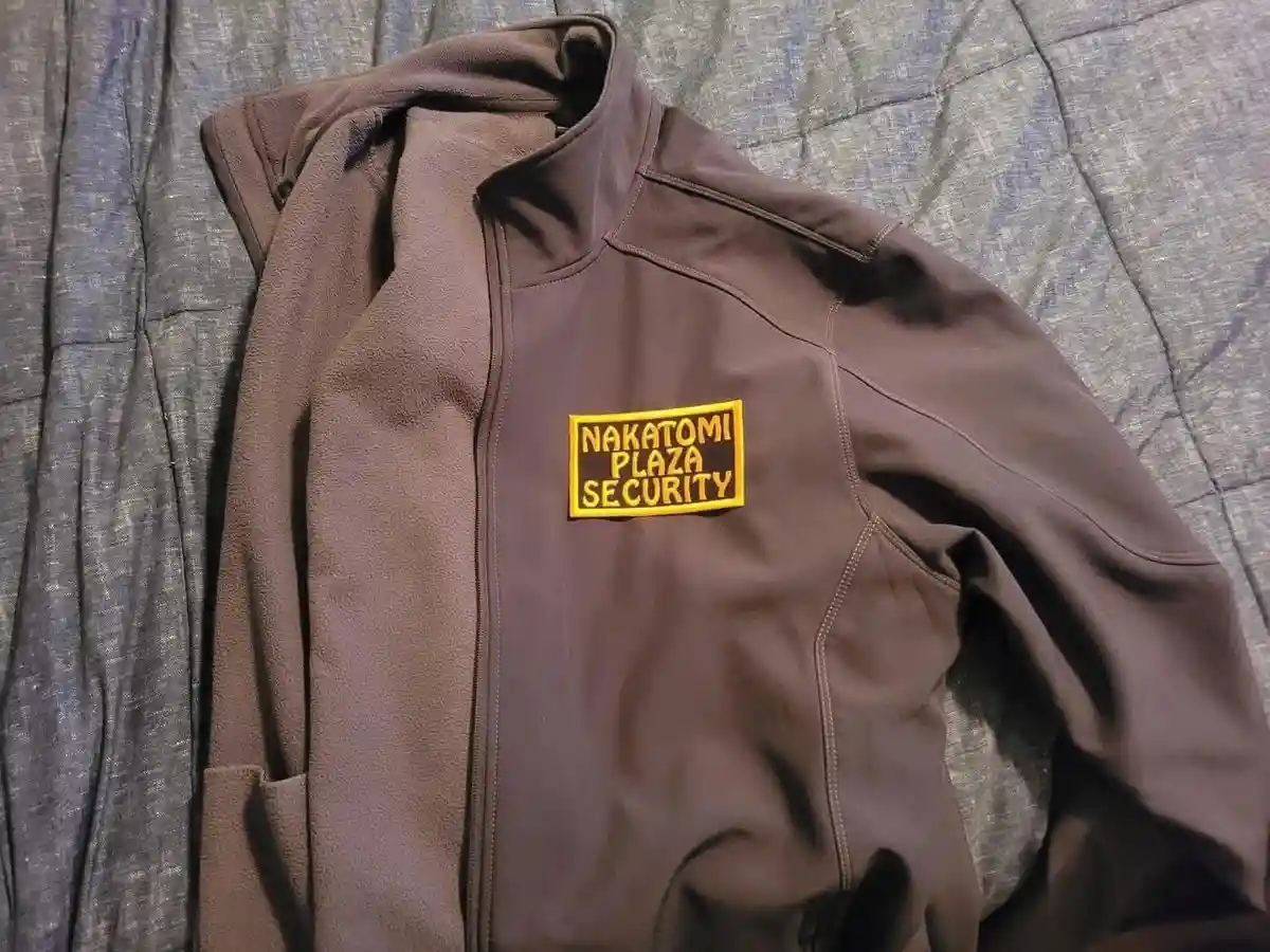 A brown zip-up fleece jacket lies on a gray textured surface, featuring a yellow rectangular patch on the left chest that…
