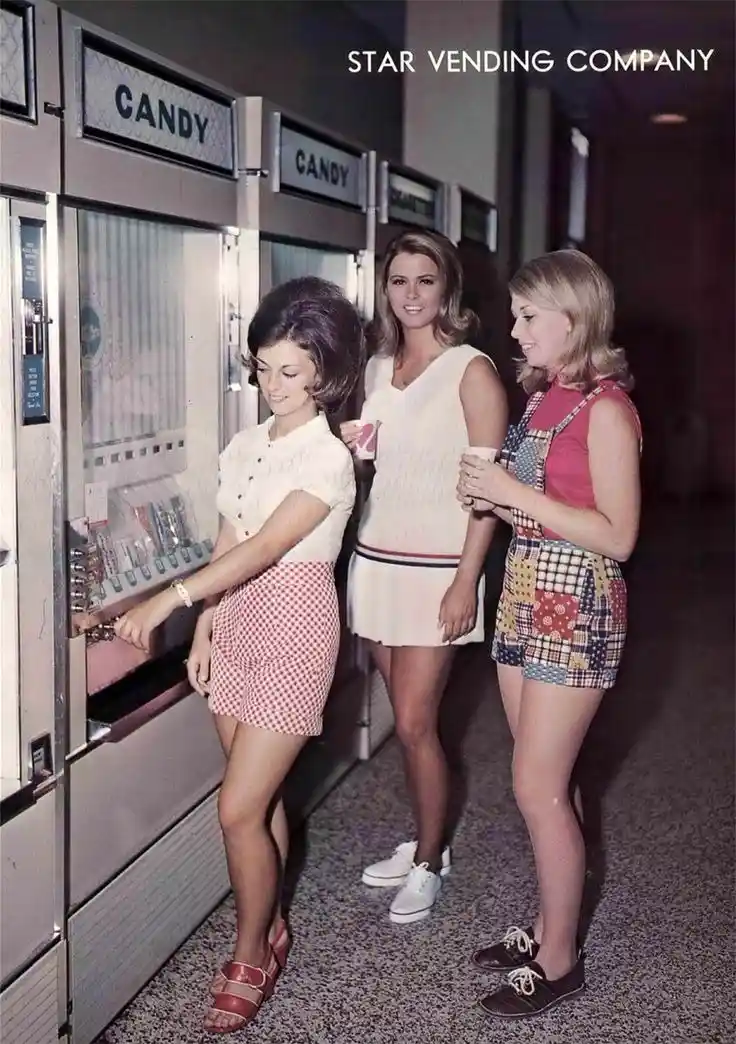 Three women in 1960s fashion stand near vintage candy vending machines labeled 'CANDY'. The woman on the left inserts money…
