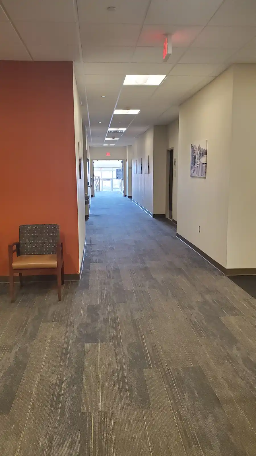 A long, empty office hallway with gray carpet, beige walls, and a single chair against an orange wall. Red exit signs are…