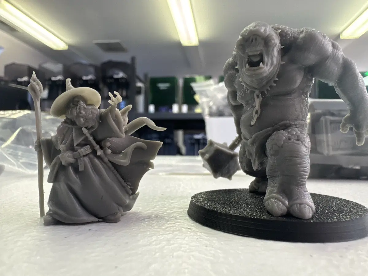 Two un-painted 3D-printed fantasy miniatures sit on a white surface: a wizard in a wide-brimmed hat and robe holding a…