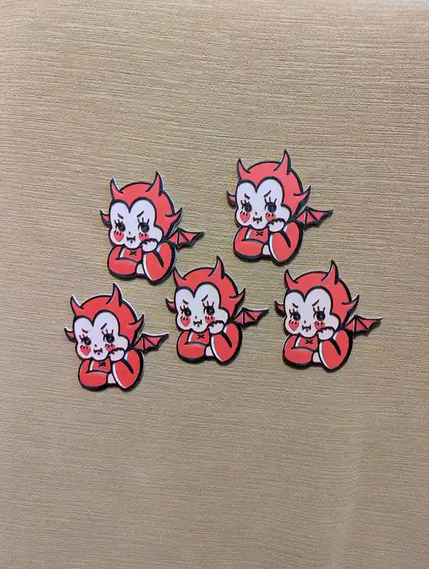 Five identical red and white cartoon devil pins with cute, blushing faces and small wings are arranged on a light brown…