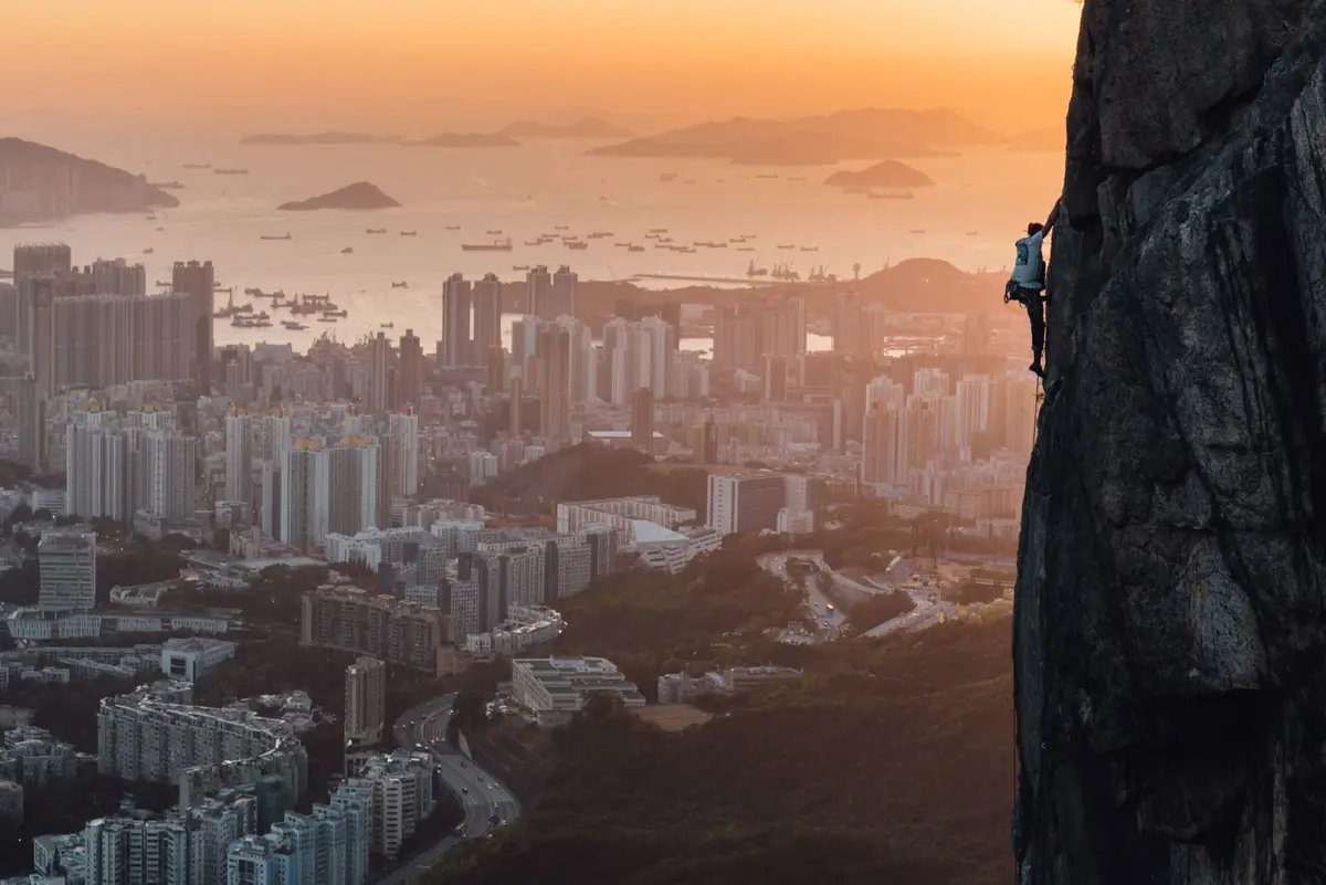 A climber in a white shirt and backpack is scaling a steep, dark rock face on the right side of the image, overlooking a…