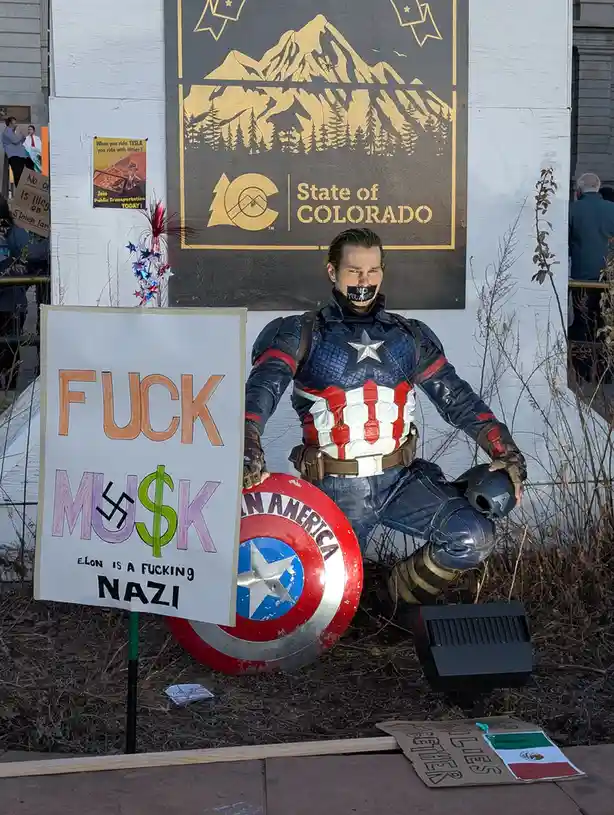 A person dressed as Captain America kneels outdoors holding his shield, with a protest sign reading 'FUCK MUS$K' and 'ELON…