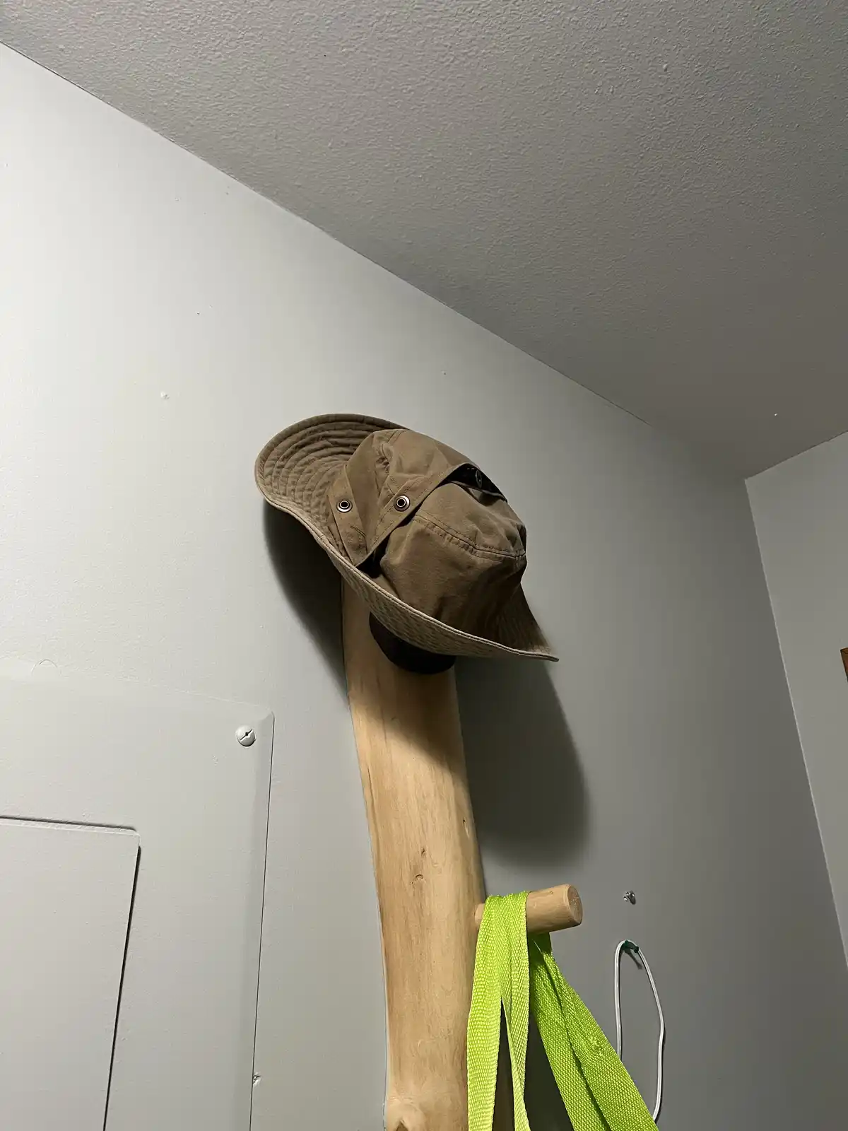 A brown bucket hat is perched atop a tall, light-colored wooden coat rack in a corner with white walls and ceiling. A bright…