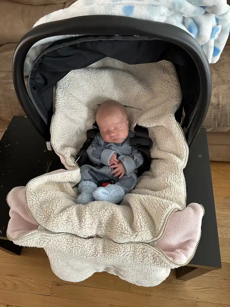 A newborn baby, dressed in a blue onesie and socks, is peacefully sleeping in a car seat that is covered with a fluffy…
