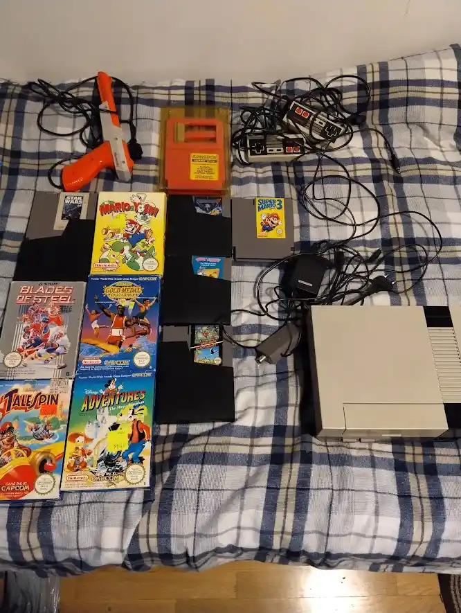 A top-down photo shows a Nintendo Entertainment System console, two controllers, a Power Pad, a Game Genie, and several game…