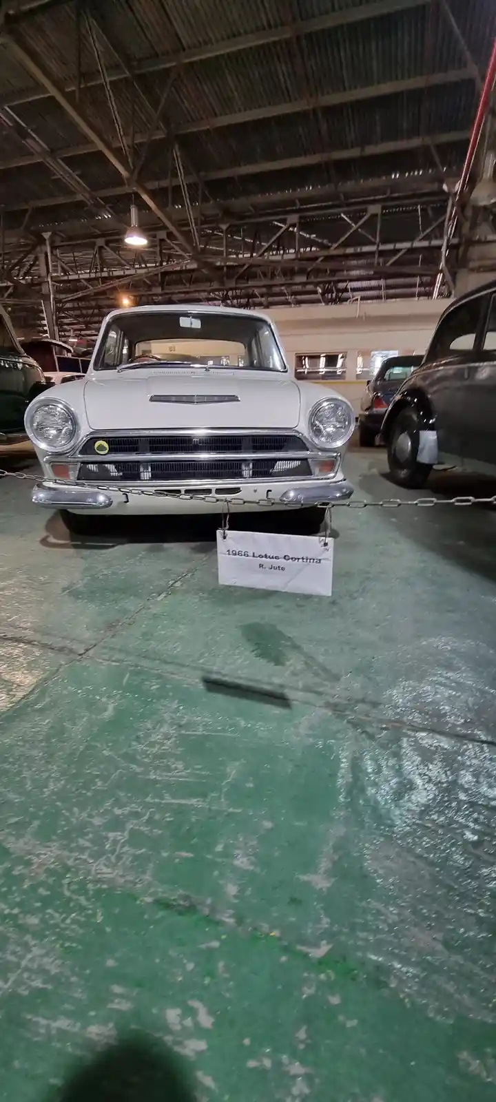 A white 1966 Lotus Cortina is displayed in a museum or garage with a green floor and metal ceiling, with a sign hanging from…