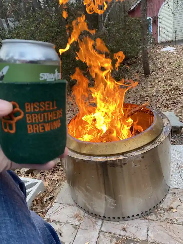 A hand holds a beer can wrapped in a green sleeve that reads 'BISSELL BROTHERS BREWING' next to a metal fire pit with bright…