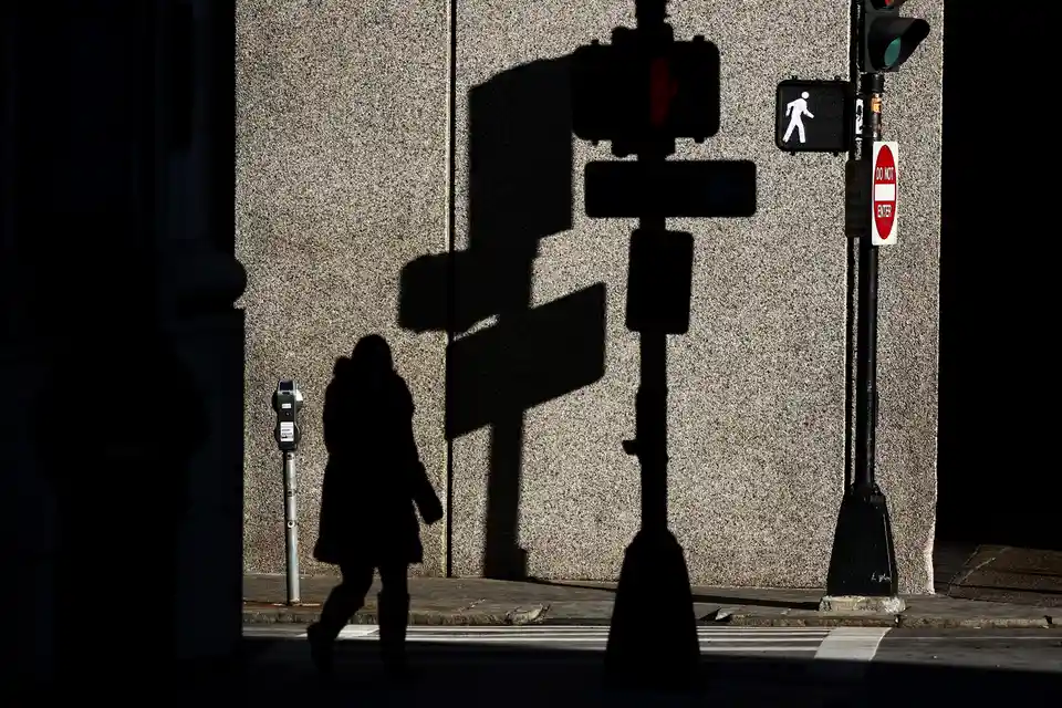 A silhouette of a person walking on a city sidewalk next to a parking meter, with the large, dark shadow of a traffic light…