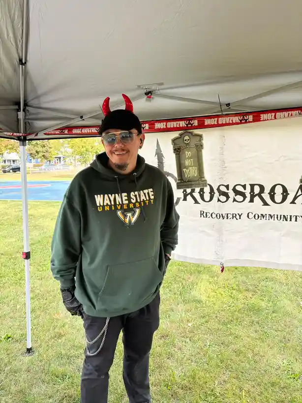 A man wearing a Wayne State University hoodie, red devil horns, sunglasses, and black gloves stands smiling under a tent…