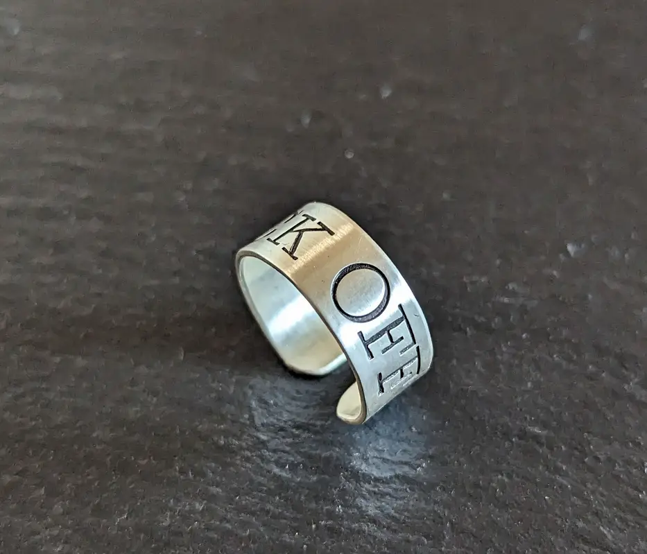 A silver-colored metal ring with the letters 'K' and 'E' and a circle engraved on its surface, resting on a dark textured…