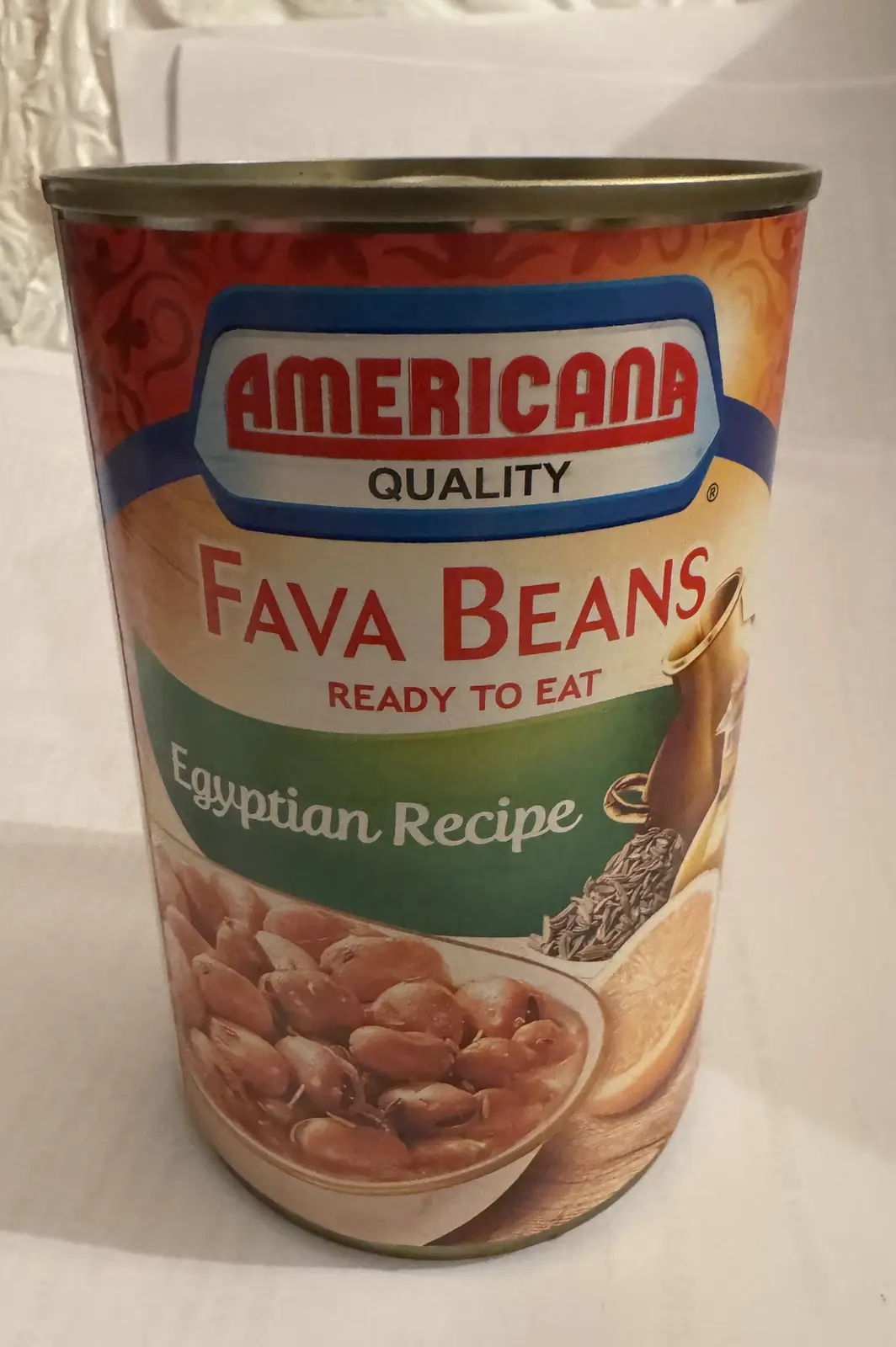 A can of Americana brand fava beans, labeled 'Ready to Eat' and 'Egyptian Recipe', sits on a white surface. The label…