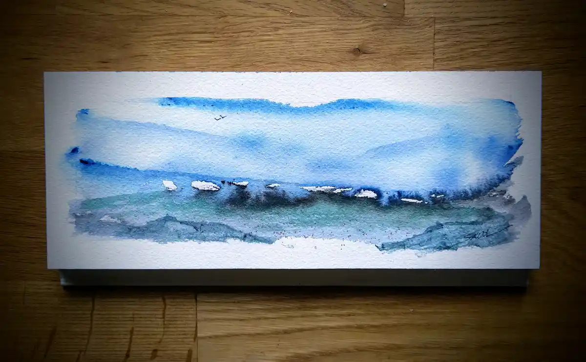 A watercolor painting on textured paper depicts a serene coastal scene with layered blues and greens, white foam at the…