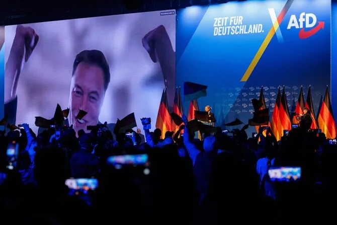 A large screen at an AfD political rally displays a smiling Elon Musk with raised fists, while a crowd holds German flags…