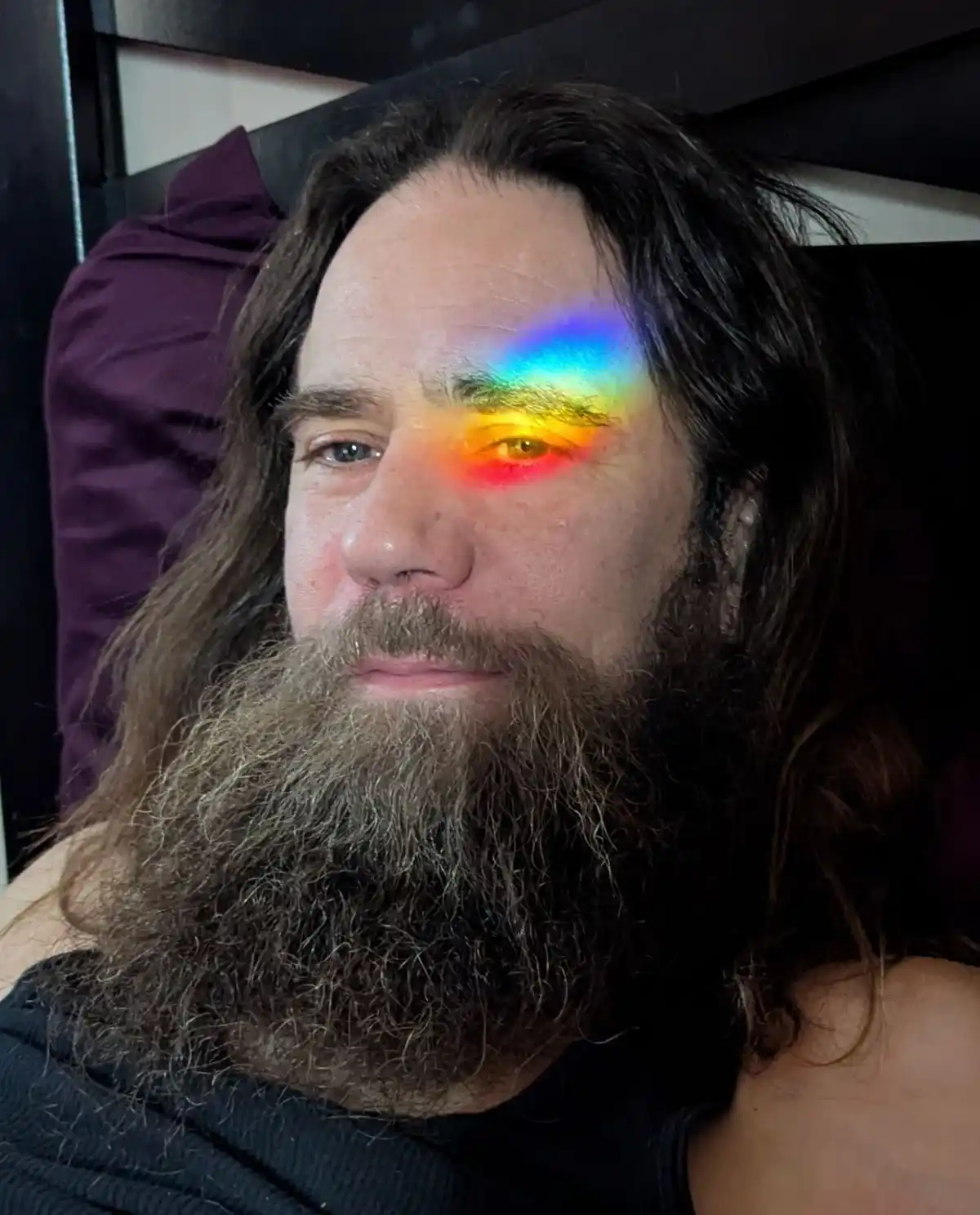 A man with long dark hair and a full beard is shown in a selfie, looking directly at the camera with a neutral expression. A…