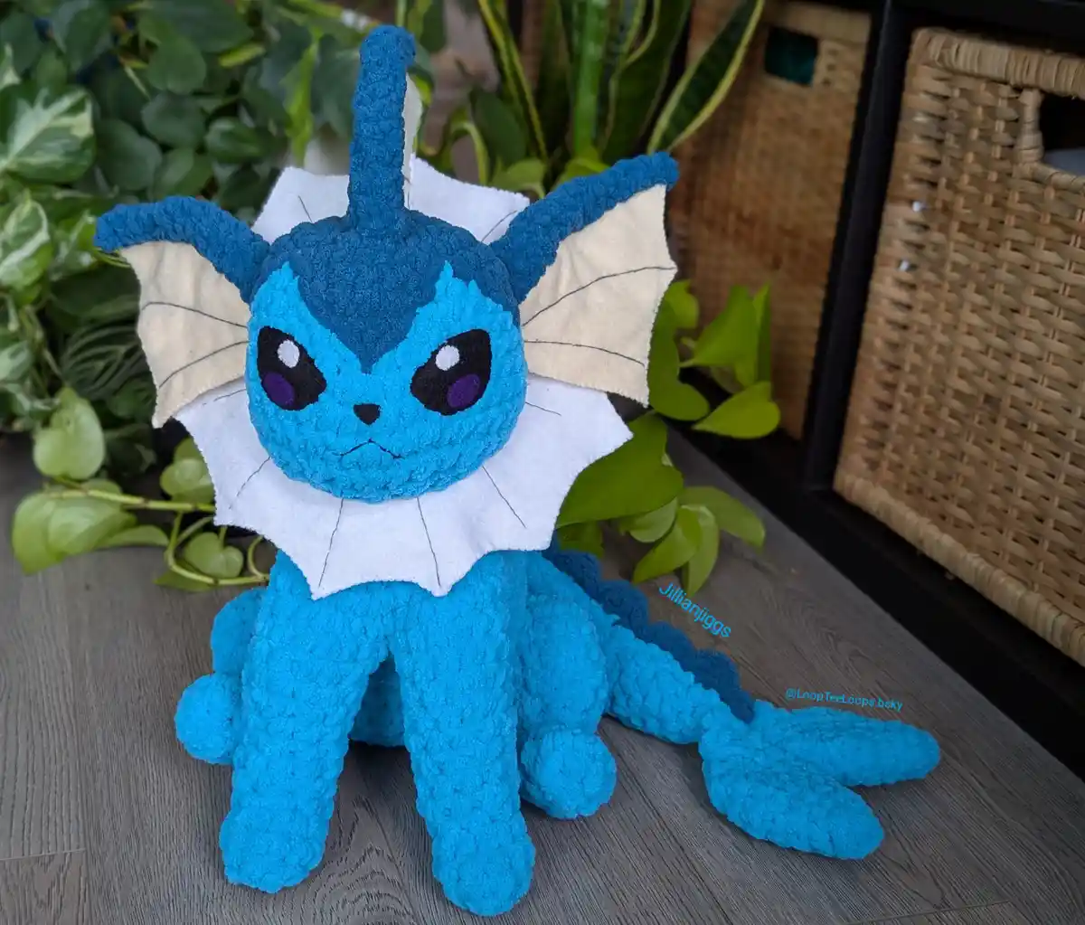 A handmade, crocheted plush toy of the Pokémon Glaceon sits on a wooden floor surrounded by green plants. The plush is…