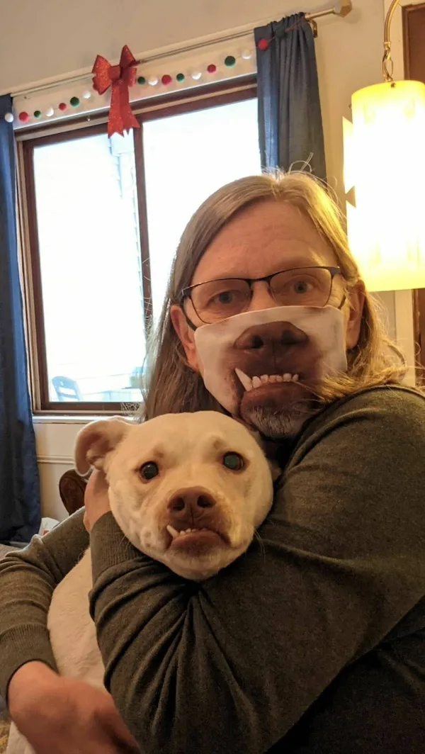 A person with long hair and glasses wears a face mask printed with a dog's face, while hugging a white dog that also has its…