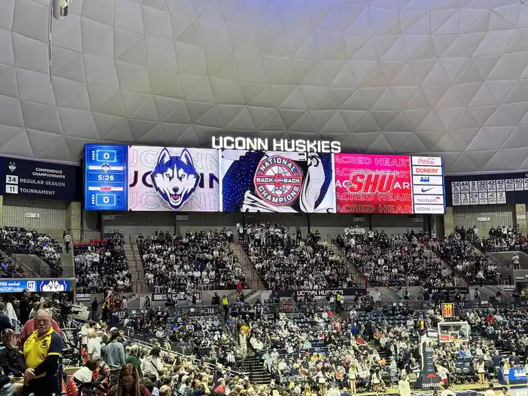 A wide shot of the UConn Huskies basketball arena shows a large scoreboard displaying 'NATIONAL CHAMPIONS' with 'BACK UCONN…