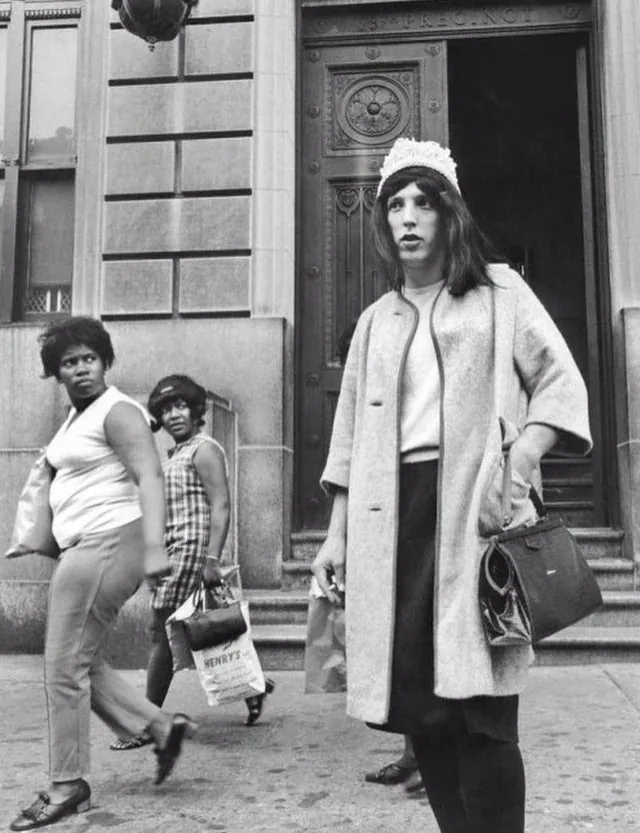 A black-and-white street photo shows a woman in a light coat and white knit hat standing in front of a building, looking…