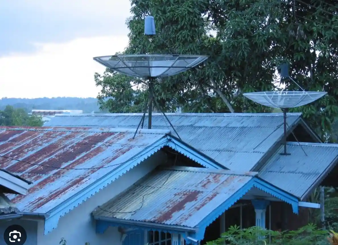 Two large, clear satellite dishes are mounted on the rusty, blue-painted corrugated metal roof of a house, with green trees…