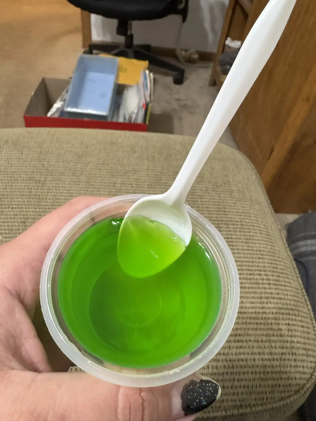 A hand holds a clear plastic cup filled with bright green, translucent gelatin, with a white plastic spoon lifting a scoop…